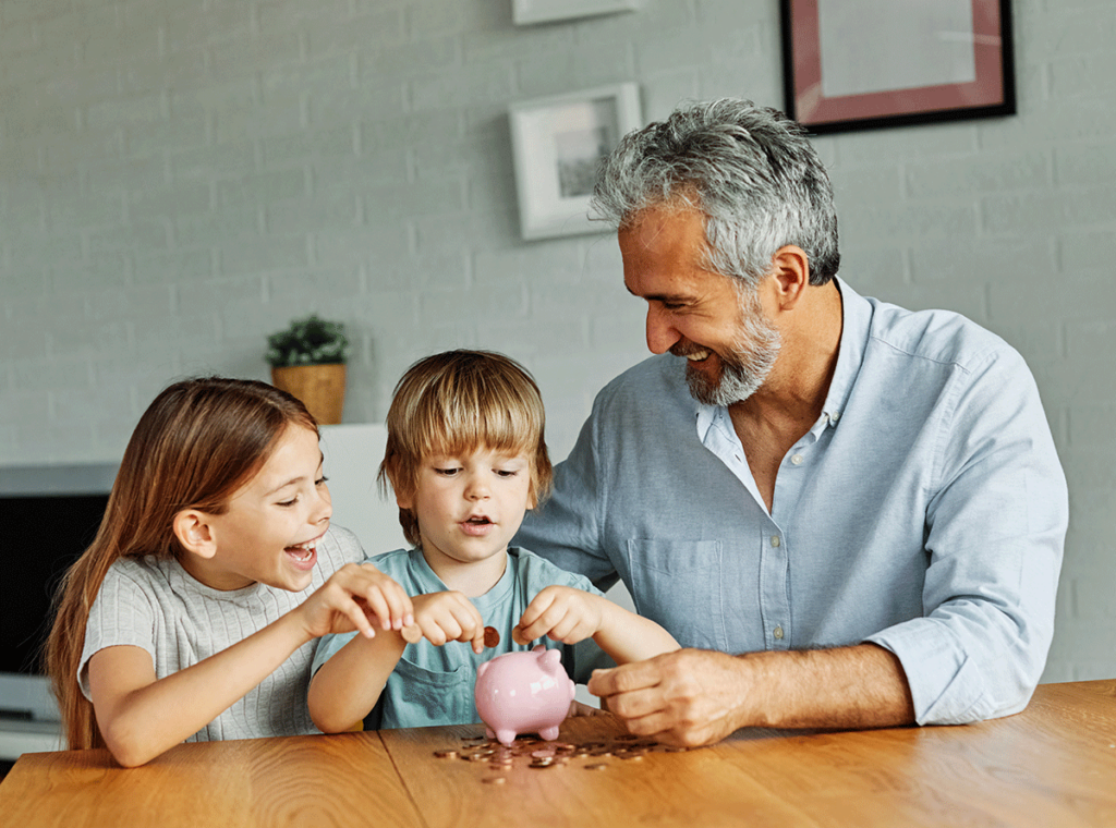 older man teaching two children how a piggy bank works as he displays a brilliant white smile